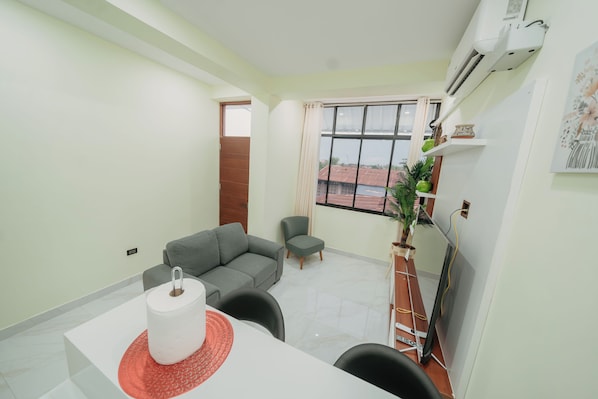A modern living room with a grey sofa, a small table with a white object, and a window with a view of buildings.