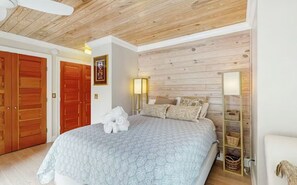 A bedroom with a bed, wooden ceiling, a wall-mounted shelf, and a door.