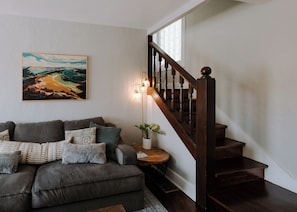 Ein Wohnzimmer mit einer dunklen Holztreppe, einem Sofa mit gemusterten Kissen, einem Gemälde an der Wand und einer Stehlampe.
