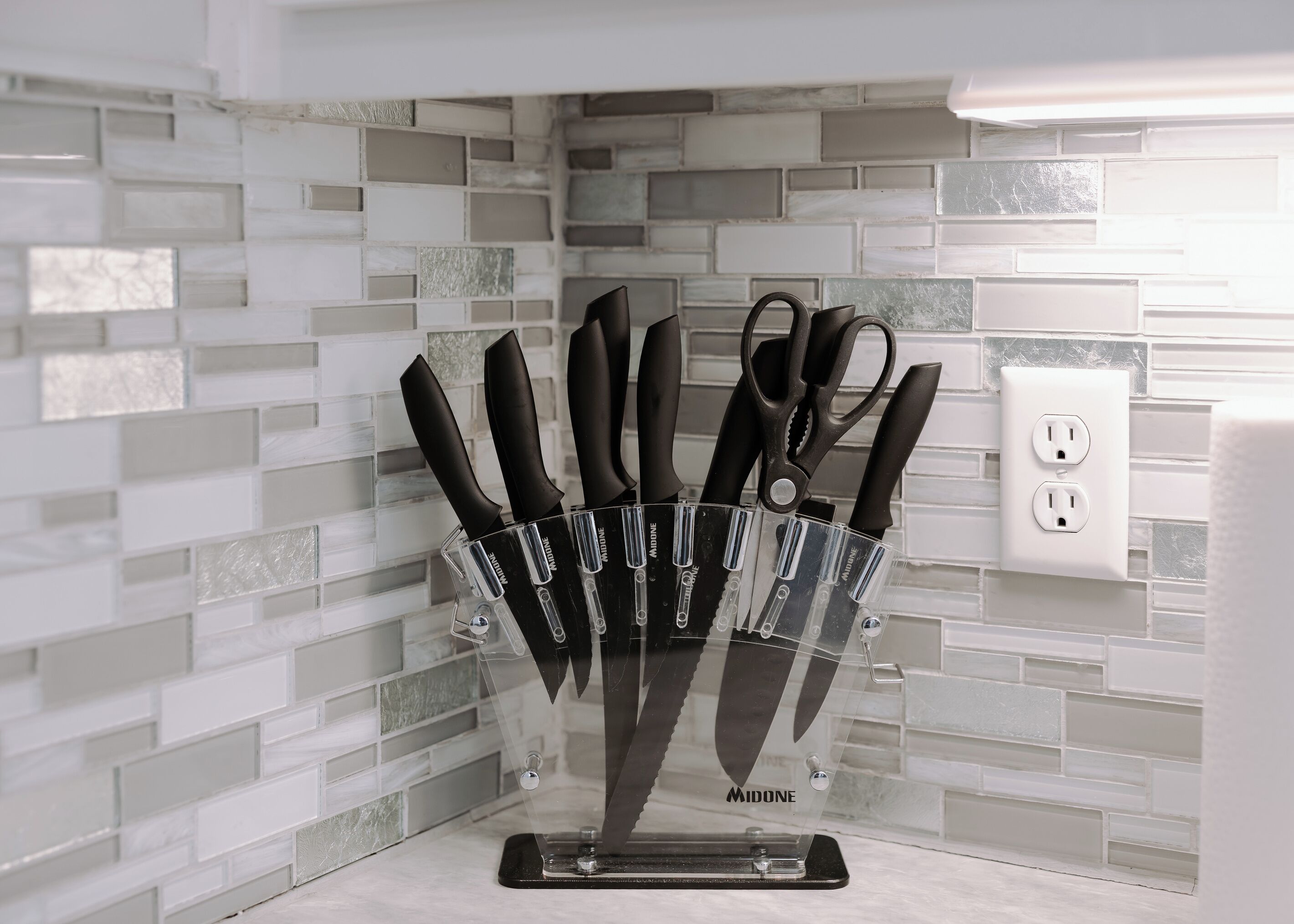A kitchen with a knife block holding various knives, a power outlet, and a tiled backsplash.