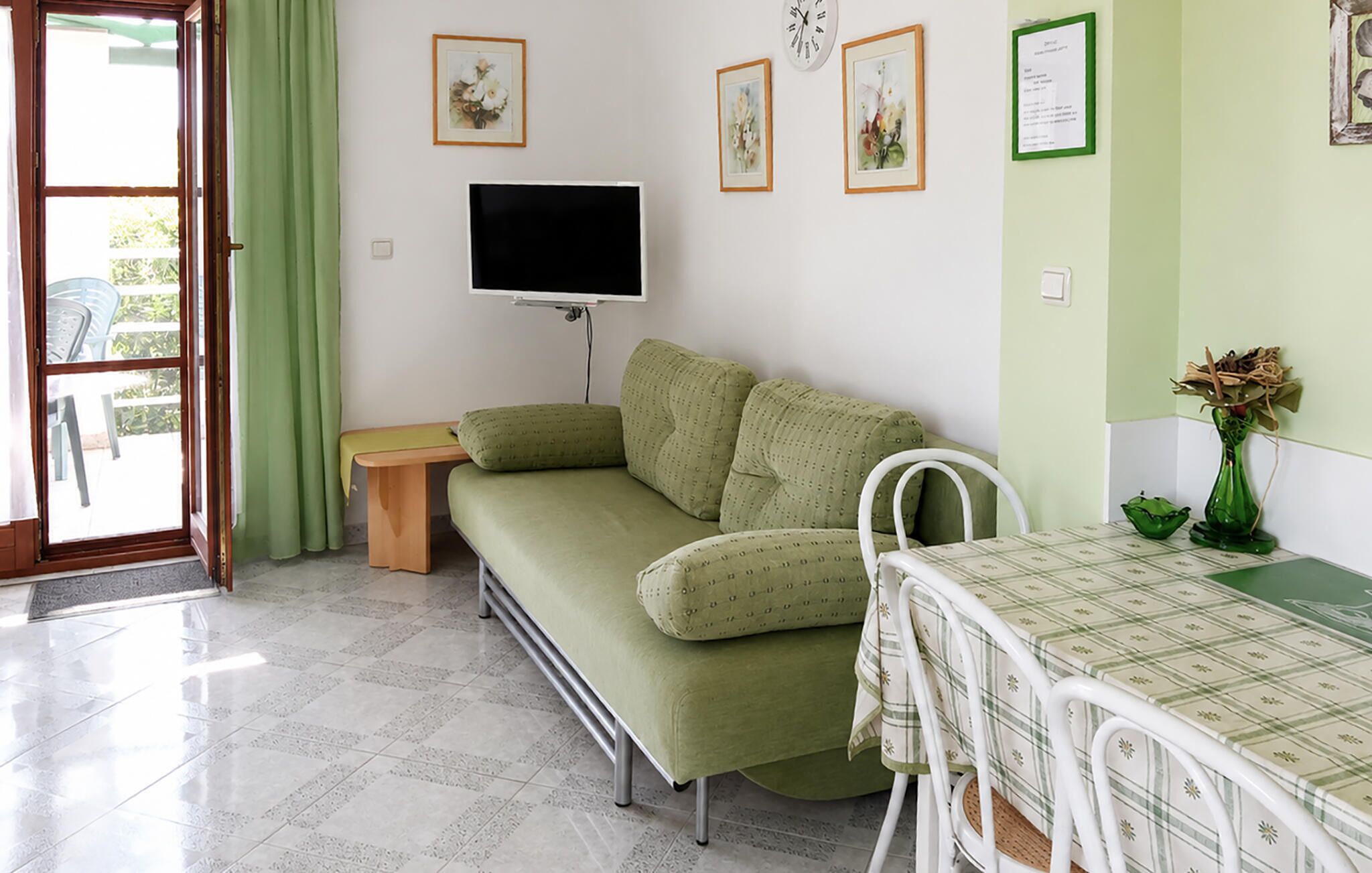 Une pièce avec un canapé vert, une petite table recouverte d’une nappe à carreaux, un téléviseur fixé au mur et une porte donnant sur l’extérieur.