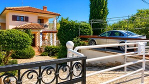 A two-story house with a tiled roof, a car parked in the driveway, and a white fence with decorative metalwork. - Lovely apartment in Kukljica with WiFi (Kukljica)