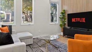 Ein modernes Wohnzimmer mit einem großen Flachbildfernseher, einem Glastisch und einer Couch mit Kissen.