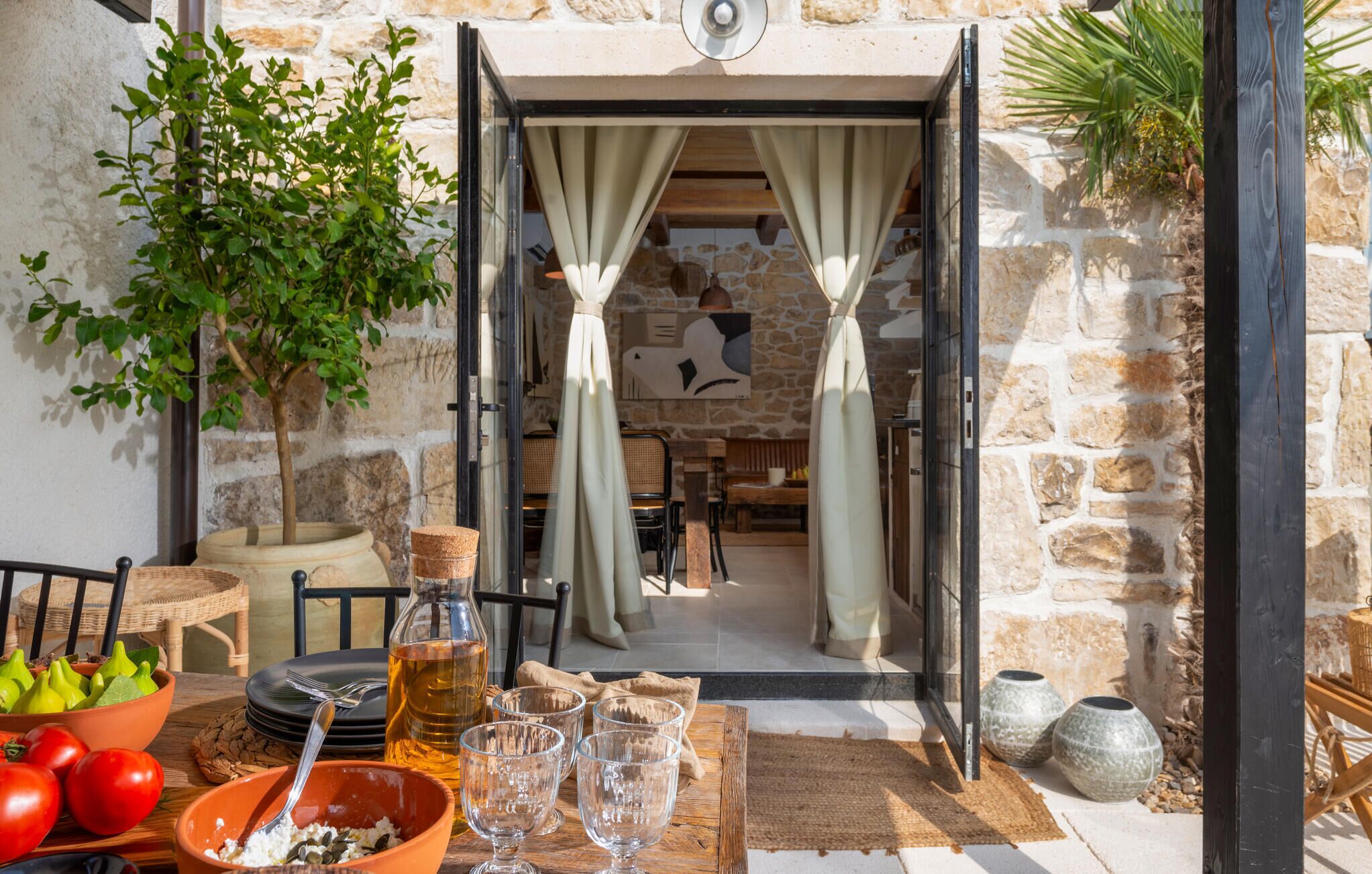 An outdoor dining area with a stone wall, a table set with plates, glasses, and a bottle, and a view into the interior.