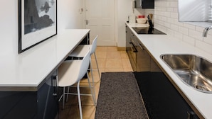 Een compacte keuken met witte kasten, een RVS spoelbak, een klein eethoekje met een witte tafel en stoelen, en een lijstwerkkunstwerk aan de wand.