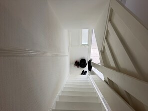 Una escalera blanca con barandilla que conduce a un rellano donde hay zapatos y una mochila.