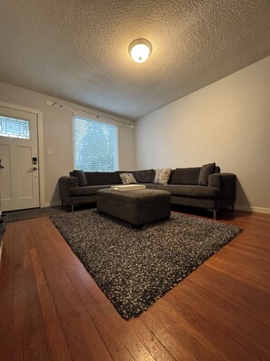 Ein Wohnzimmer mit einem Ecksofa, einem Hocker, einem gemusterten Teppich und Holzdielen.