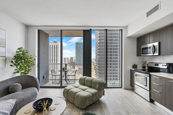 A modern apartment with a sofa, a dining table, a kitchen with a microwave, and a large sliding glass door leading to a balcony. - Luxury High-Rise Bay Views + Pool + Parking Steps to Bayside & Brickell (Miami)