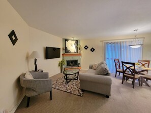 Ein Wohnzimmer mit Sofa, Sessel, Fernseher, Kamin und Essbereich mit Stühlen und Tisch.