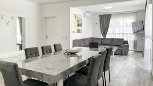 Ein modernes Wohnzimmer mit Essbereich, grauen Sofas, einem großen Esstisch und einem großen Fenster mit Vorhängen.