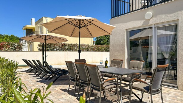 Eine Terrasse im Freien mit Tisch, Stühlen und Sonnenschirmen, mit Blick auf ein Gebäude.
