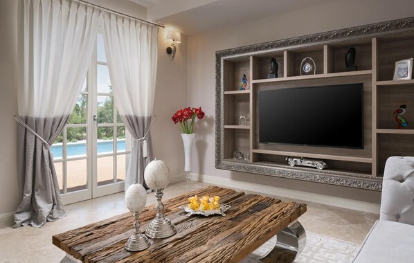 Ein Wohnzimmer mit großem Fernseher, Couchtisch aus Holz und Blick auf einen Pool.