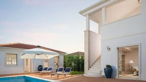 Uma área de piscina com espreguiçadeiras e guarda-sóis, um edifício branco com escadas e uma grande planta em vaso.