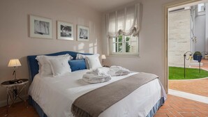 Quarto com cama, mesa de cabeceira, luminária, janela com cortinas e quadros nas paredes.