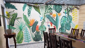 Comedor con un mural de plantas tropicales, una mesa de madera, sillas y una planta en maceta sobre una mesita.