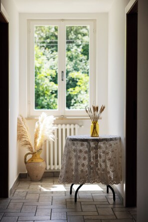 Ein Zimmer mit Fenster, einem Tisch mit Spitze-Tuch und einer Vase mit getrockneten Pflanzen.