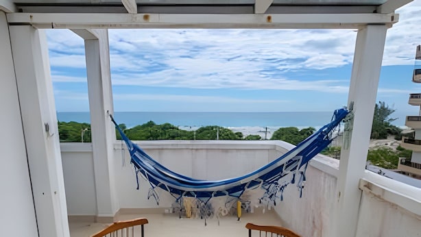 A balcony with a wooden table and chairs, a blue hammock, and a view of the sea. - Seu Lar na Praia Grande (Arraial do Cabo)