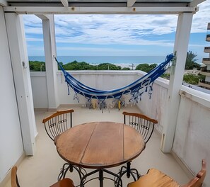 A balcony with a wooden table and chairs, a blue hammock, and a view of the sea. - Seu Lar na Praia Grande (Arraial do Cabo)