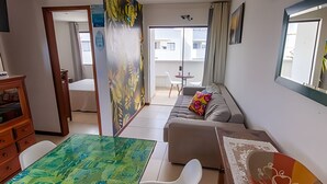 A room with a sofa, a dining table, a chair, a small table, and a view through a glass door to an outdoor balcony. - Seu Lar na Praia Grande (Arraial do Cabo)
