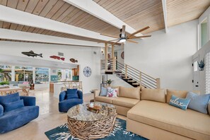 Ein geräumiger Wohnbereich mit hoher Decke, einem Glastisch, einem beigen Sofa und einem blauen Sessel.