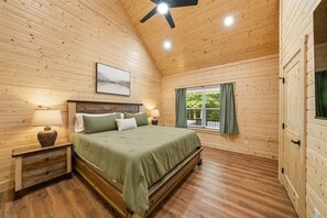 A wooden-paneled bedroom with a bed, bedside lamps, a ceiling fan, and a window with curtains. - Modern, Cozy Cabin Close to Major Attractions! (Pigeon Forge)