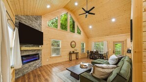 L’intérieur chaleureux d’un chalet avec une cheminée, des murs en bois, un coin repas et un ventilateur de plafond.