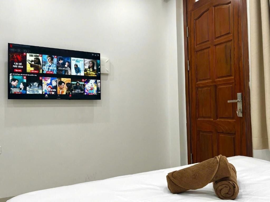 A hotel room with a wall-mounted TV displaying various shows, a wooden door, and a neatly made bed with a rolled towel.