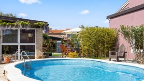 Piscina con borde de piedra, terraza de madera y zona ajardinada con plantas y una silla.