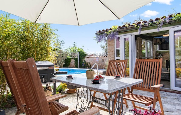 An outdoor patio with a pool, wooden chairs, a table, and a grill.