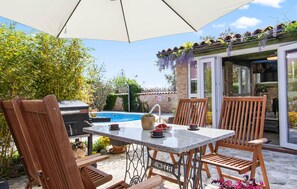 An outdoor patio with a pool, wooden chairs, a table, and a grill.