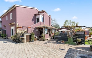 A pink building with a tiled roof, a covered porch, and a paved courtyard with potted plants.