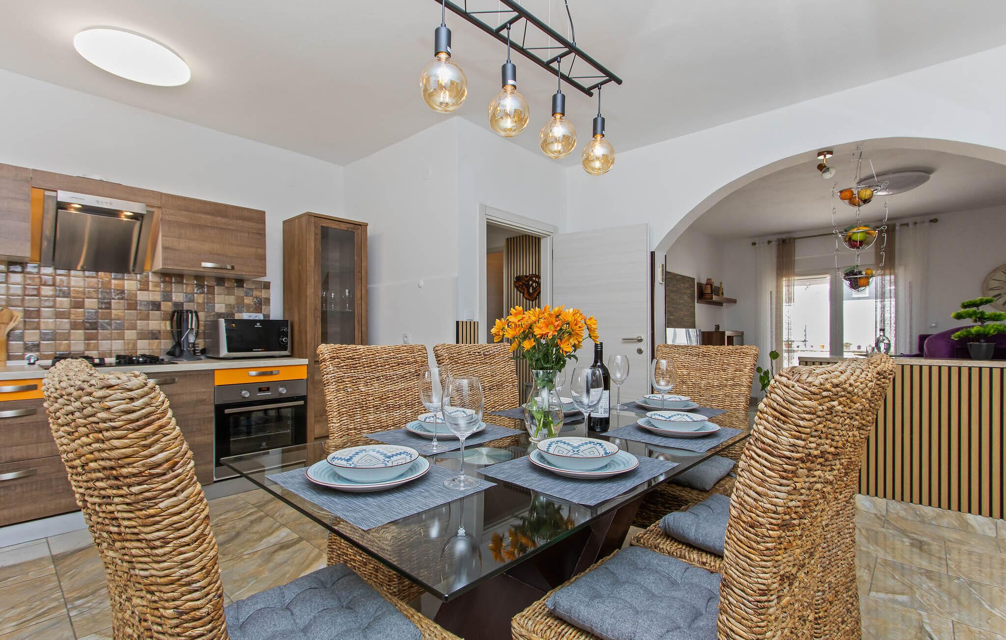 A modern kitchen with a dining area, featuring a round table with chairs, a vase with flowers, and a hanging light fixture.