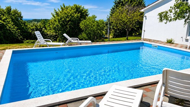 Une piscine rectangulaire à l’eau d’un bleu cristallin, entourée de chaises longues blanches et d’un bâtiment blanc doté d’un toit recouvert de tuiles.