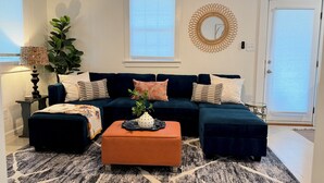 Een woonkamer met een donkerblauwe hoekbank, een oranje poef, een kamerkleed met patroon, een grote plant en een decoratieve spiegel aan de muur.