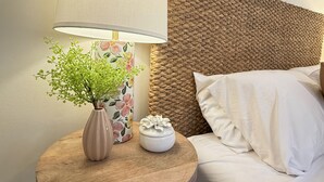 Een bed met witte beddengoed, een rond houten nachtkastje met een vaas en een lamp, en een geweven hoofdbord.