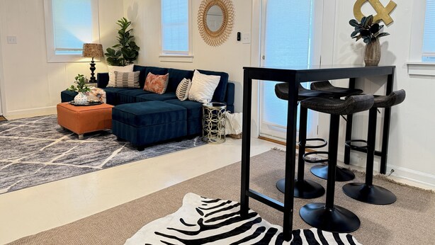 Een woonkamer met een donkerblauwe hoekbank, een vloerkleed met zebraprint, een plafondventilator en een eethoek met zwarte barkrukken.