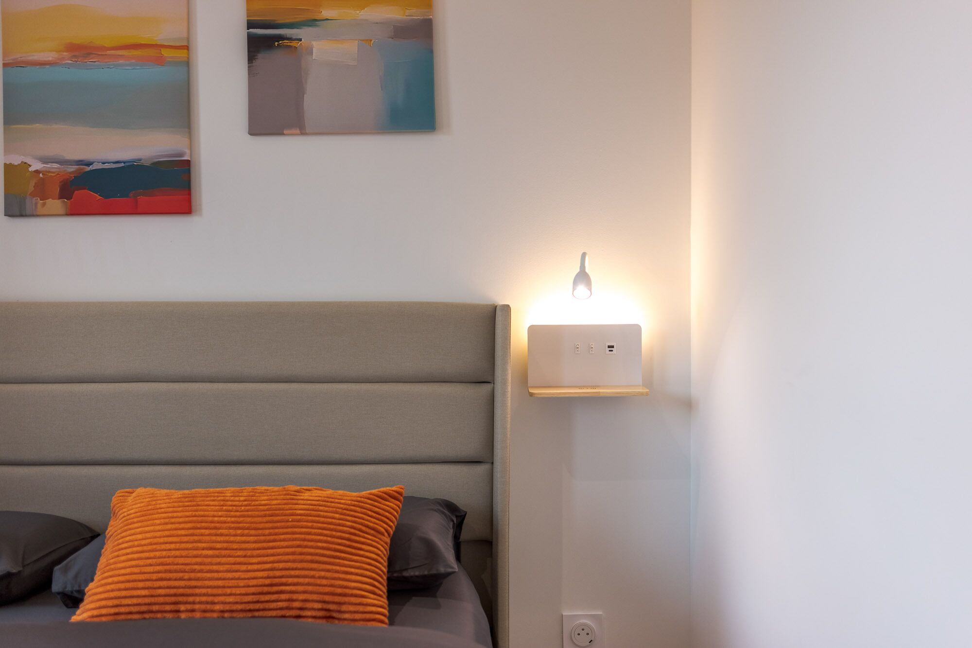 Uma cama com cabeceira cinza, uma luminária fixada na parede e duas pinturas na parede.