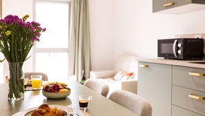 Ein Frühstückstisch mit einem Croissant, Obstschale und Kaffee, dazu eine Blumenvase und Blick in ein Wohnzimmer.