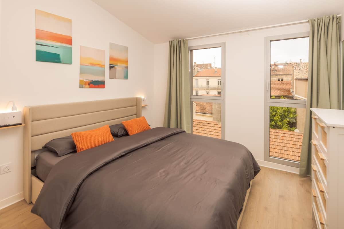 Un dormitorio con cama, mesita de noche, ventana con cortinas y vistas a edificios.