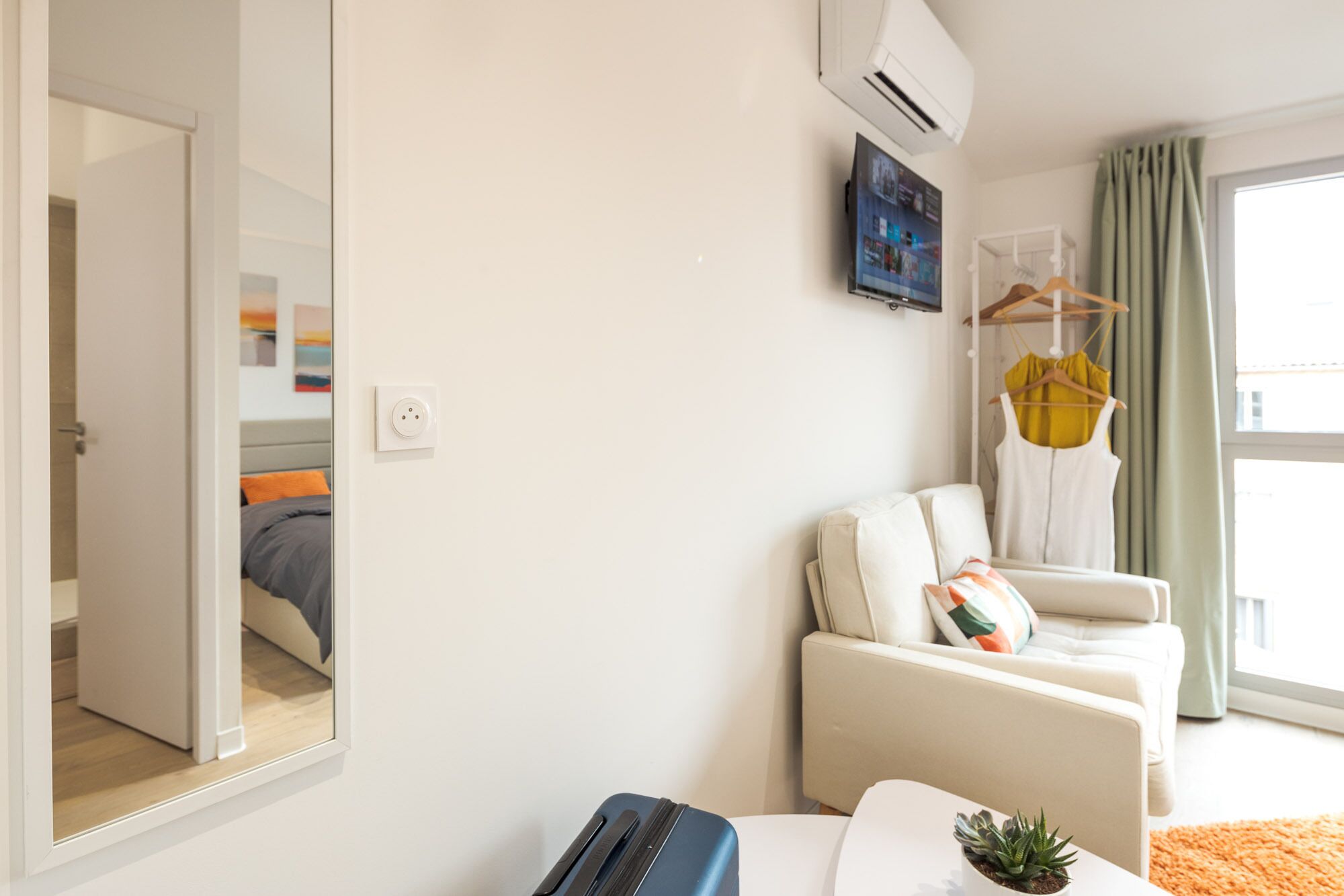 Une chambre d’hôtel moderne comprenant un lit, un coin salon avec un canapé et une petite table, un miroir et une fenêtre avec des rideaux.