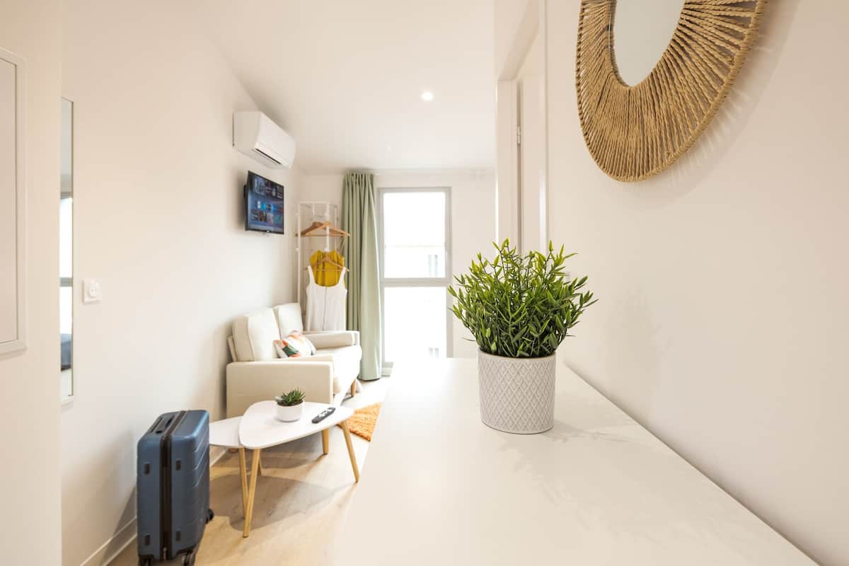 Una habitación de hotel moderna con un sofá, una pequeña mesa, una planta en maceta, una maleta y un espejo en la pared.