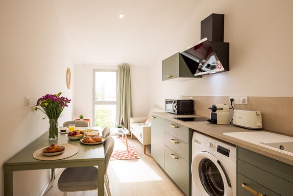 A compact kitchen with a washing machine, microwave, and a dining area with a table set for breakfast. - Maison Flaugergues (Montpellier)