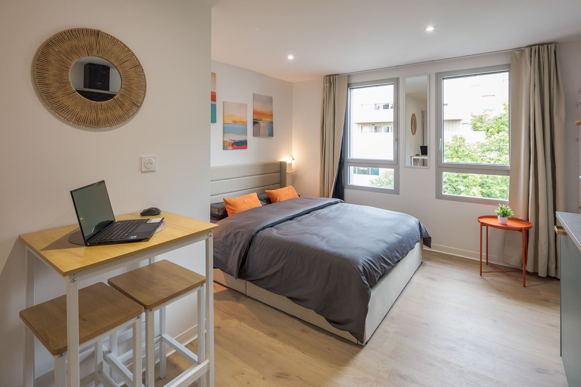 A bedroom with a bed, a desk with a laptop, a window with curtains, and artwork on the wall.