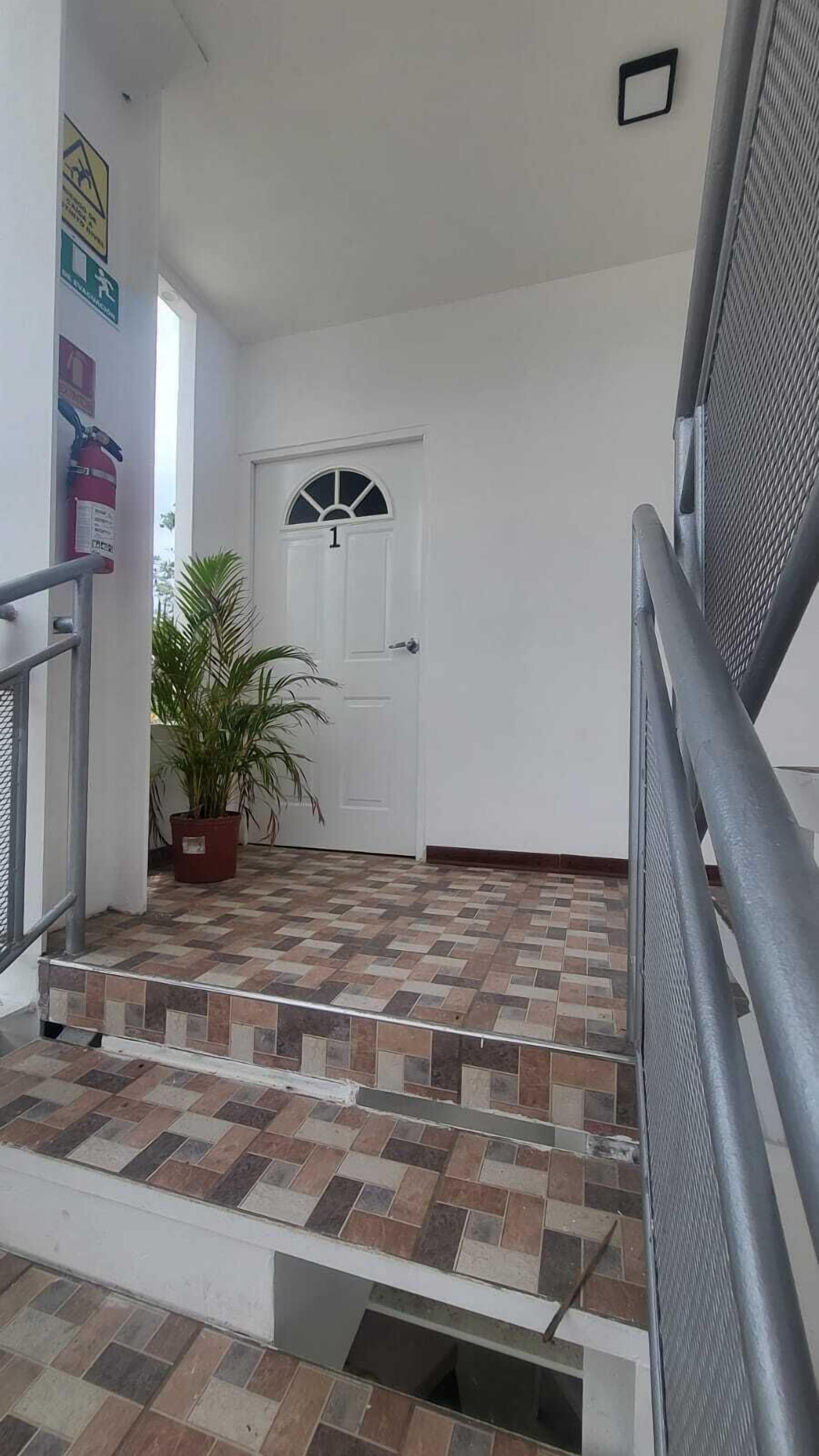 Una escalera lleva a una puerta con el número '1' encima, un extintor en la pared y una planta en maceta.