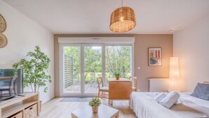 A modern room with a TV, a dining table, a sofa, a lamp, and a view through a glass door to an outdoor area. - Jardins de Flore - Studio Apartment, Sleeps 2 (Mérignac)