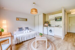 Un dormitorio pequeño con cama, mesita de noche y una pequeña mesa con una planta. Junto a él, una kitchenette con microondas, horno y lavadora.