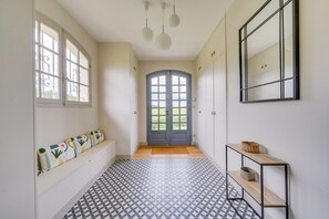 Ein Flur mit gemustertem Boden, einer Bank mit Kissen, einer Tür mit Fußmatte und einem an der Wand befestigten Regal mit Korb.