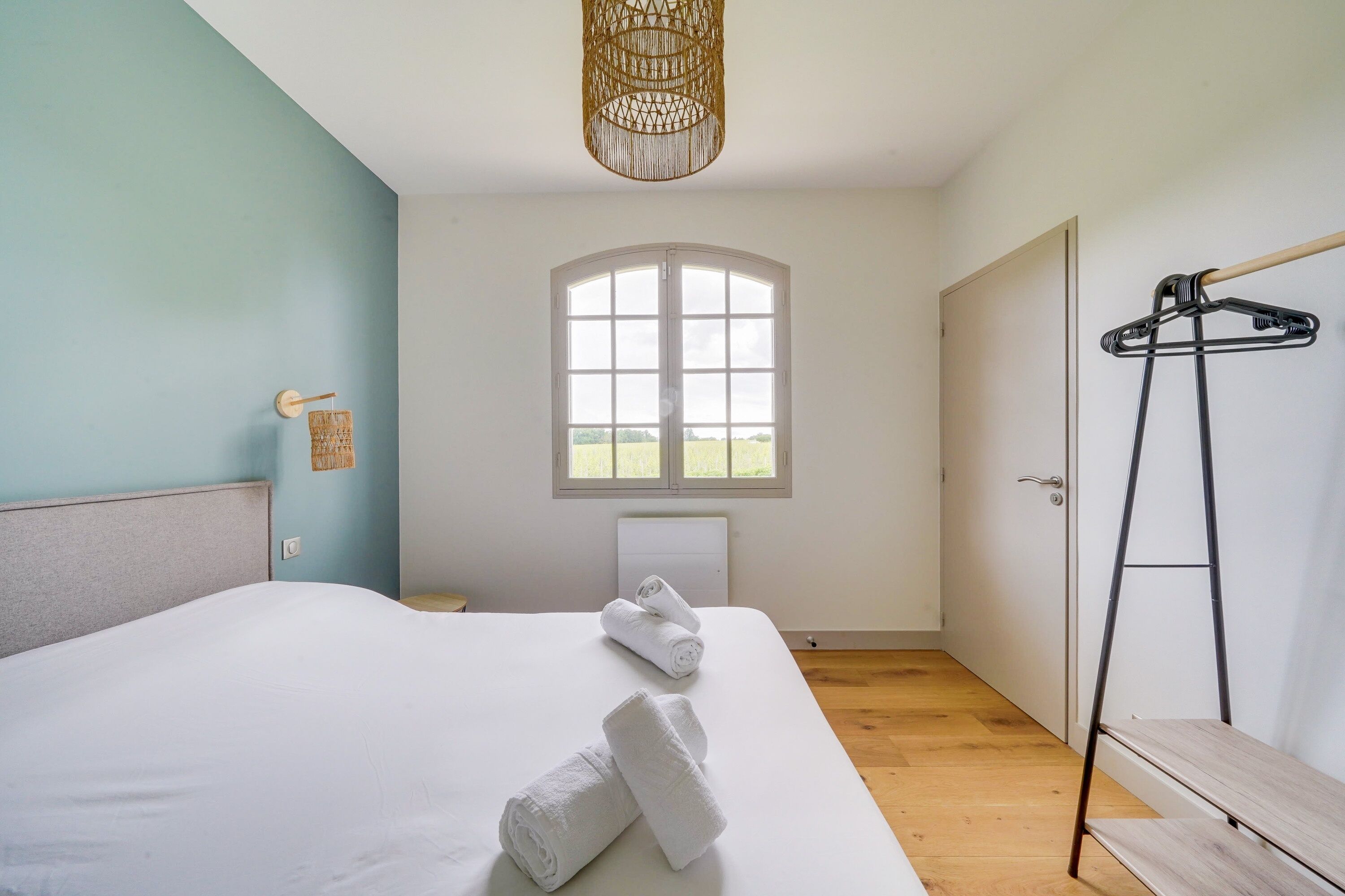 A bedroom with a bed, a window, a hanging light fixture, and a wooden floor.