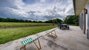 Een terras met buitenmeubilair, een wijngaard op de achtergrond en een gebouw met een overdekte doorgang.