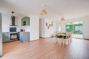Una cocina moderna con horno empotrado, microondas y armarios verdes. Hay una zona de comedor con una mesa y sillas, y un amplio ventanal corredero que da a un jardín.